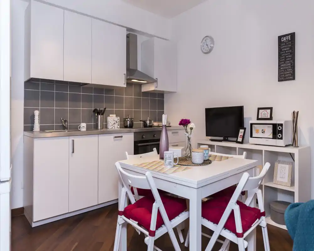 Cocina moderna y compacta con pequeña mesa de comedor. Mobiliario blanco, salpicadero gris y una mesa acogedora con cojines rojos y detalles decorativos.