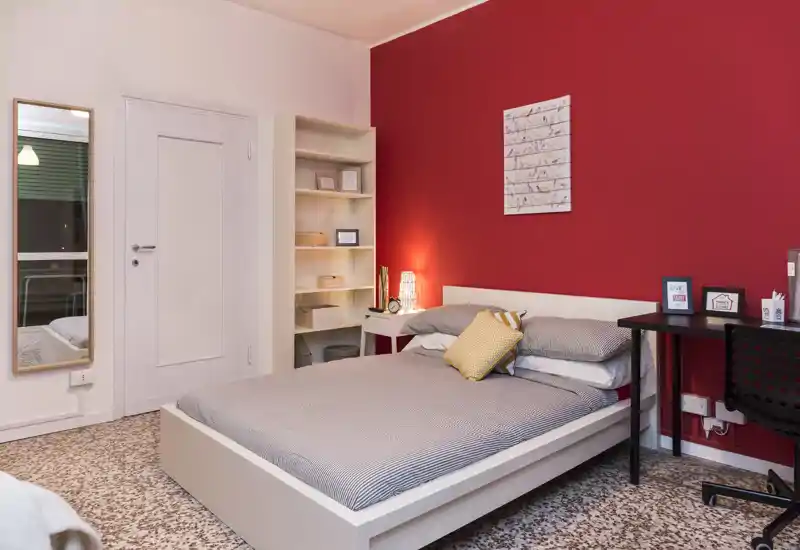 Chambre double bien aménagée avec un lit à cadre blanc contre un mur rouge, lampe de chevet, étagères et un petit bureau — espace lumineux et chaleureux, idéal pour une personne ou un couple.