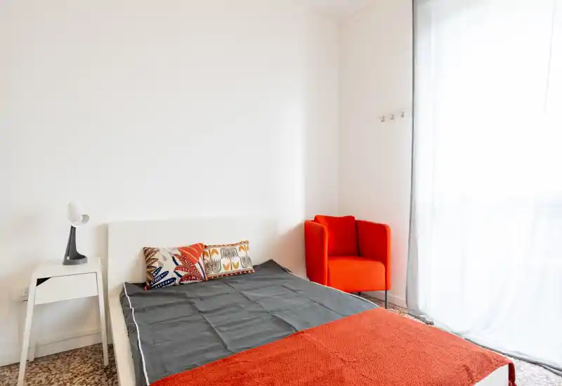 Camera da letto luminosa e minimalista con letto matrimoniale, cuscini decorativi e una sedia arancione vivace vicino alla finestra che crea un piacevole contrasto.