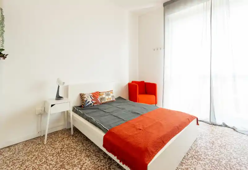 Chambre lumineuse et minimaliste avec un cadre de lit blanc, coussins colorés et plaid orange; grande fenêtre voilée apportant beaucoup de lumière et un fauteuil rouge dans un coin.