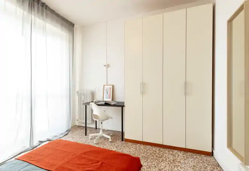 Camera da letto luminosa e minimal con un grande armadio, scrivania e una coperta arancione sul letto; molta luce naturale filtrata dalle tende leggere.
