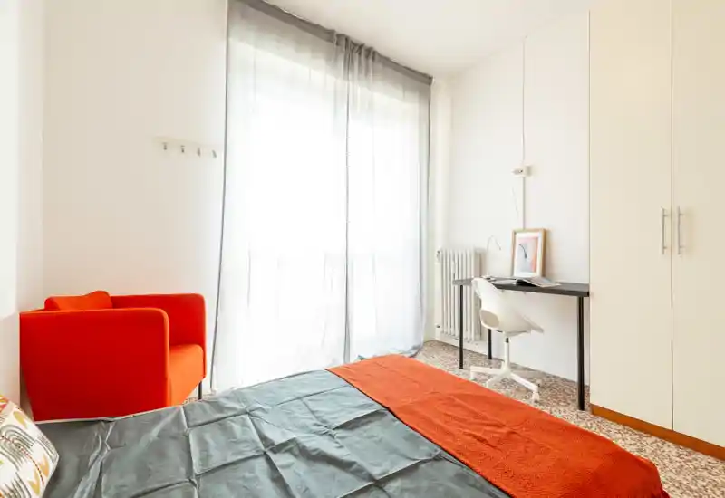 Chambre lumineuse et bien organisée avec une grande fenêtre, lit habillé de linge foncé et un plaid orange, fauteuil assorti et un petit bureau près de la penderie.
