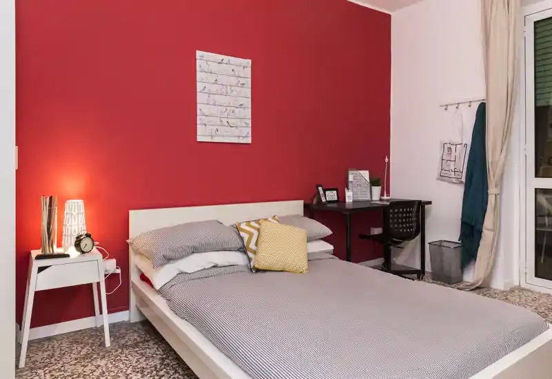Chambre lumineuse et bien mise en scène avec un lit double devant un mur rouge, table de chevet avec lampe et un petit bureau près de la fenêtre — ambiance cosy et moderne.