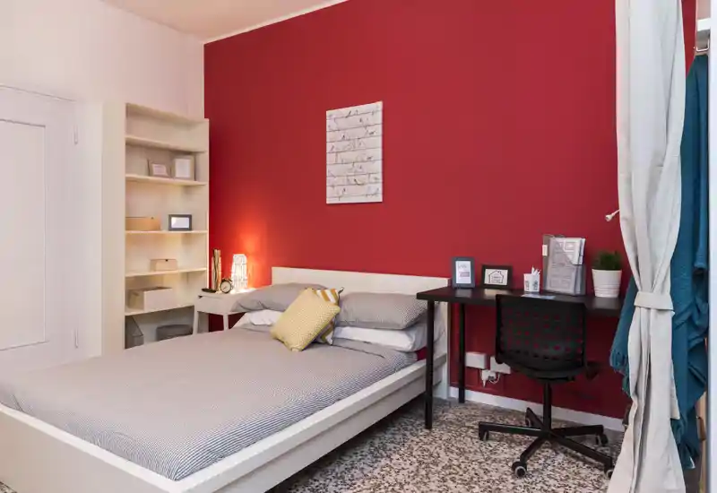 Chambre lumineuse et soignée avec un lit double blanc, linge de lit doux et un bureau devant un mur d'accent rouge vif — idéale pour une personne ou un couple.