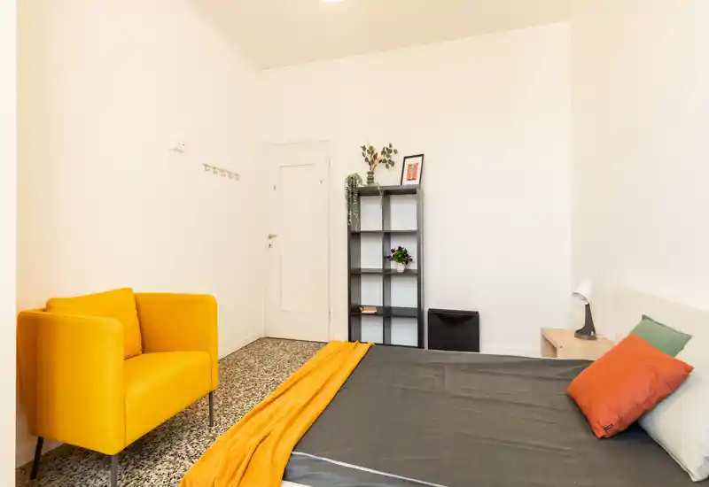 Camera da letto luminosa e ben arredata con letto matrimoniale visibile, comodino e una poltrona gialla vivace. Pareti bianche pulite, cuscini decorativi e un caldo plaid arancione che donano carattere.