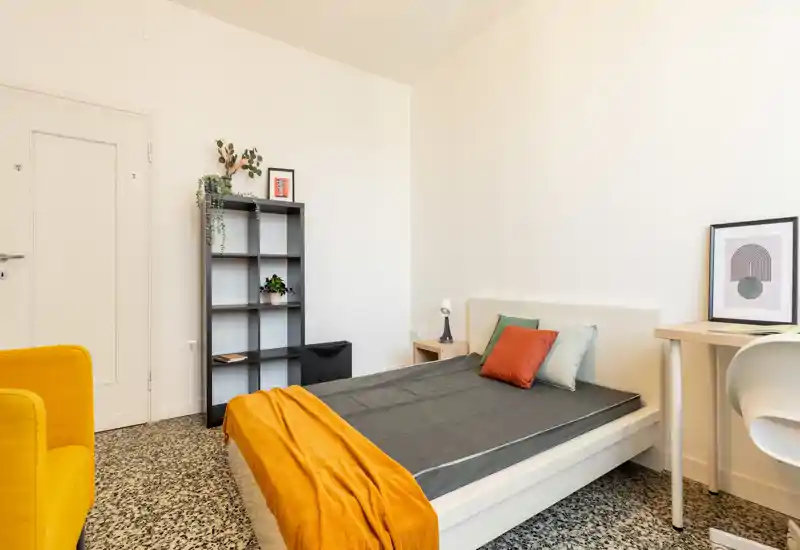 Camera da letto moderna e luminosa con letto matrimoniale ben rifatto, coperta e cuscini colorati, scrivania e libreria — perfetta per un soggiorno confortevole.