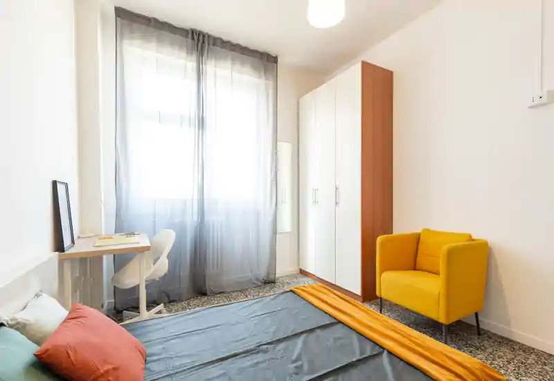 Camera da letto moderna e luminosa con letto ordinato, tende leggere alla finestra, una scrivania compatta e una poltrona gialla come elemento d’accento — perfetta per una persona.