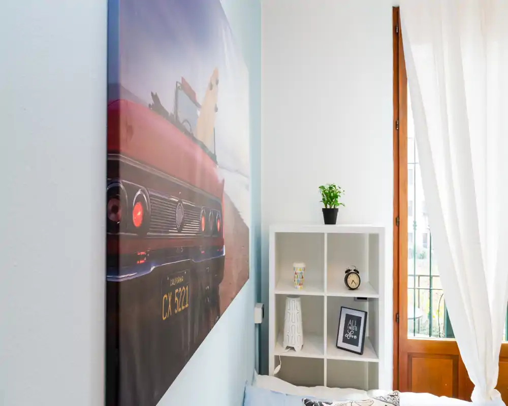 Angolo di camera da letto luminoso e raccolto con parte del letto in primo piano, una scaffalatura bianca con oggetti decorativi e una pianta, illuminato da una finestra con tende leggere.