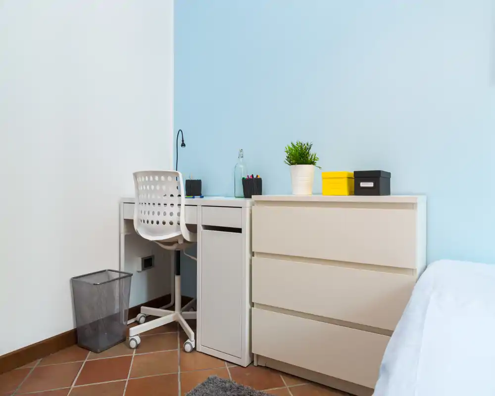 Angolo camera luminoso e minimale con scrivania bianca, cassetti e una piccola porzione visibile del letto; zona studio ordinata con pianta e accessori.