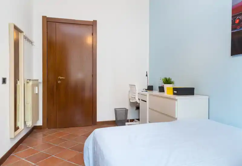 Camera semplice e pulita con letto singolo, scrivania e comò accanto a una parete azzurra chiara; ambiente luminoso e ordinato adatto a studenti o viaggiatori.