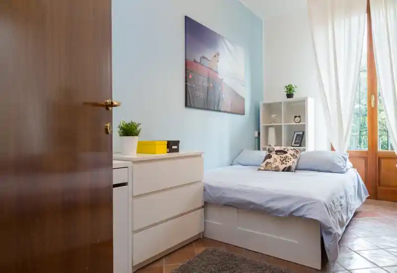 Camera da letto luminosa e ordinata con letto matrimoniale, comò bianco, scaffale e una grande finestra che lascia entrare luce naturale.