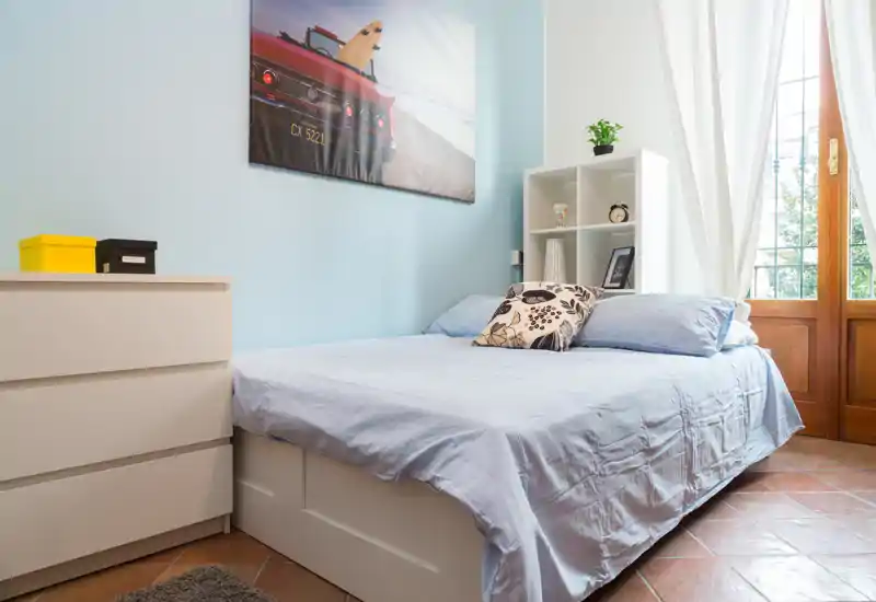 Camera da letto luminosa e ordinata con letto matrimoniale, mensole, comò e luce naturale che entra dalla grande finestra/porta.
