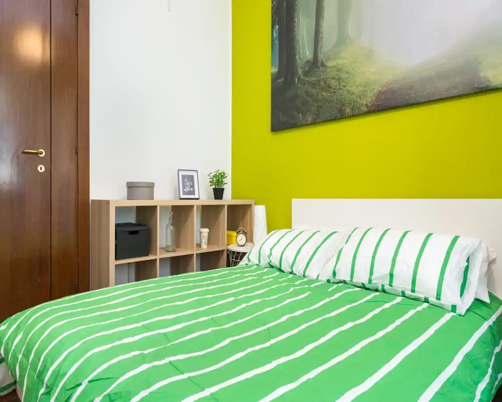 Camera da letto moderna e luminosa con copriletto a righe verdi, testiera semplice e mensole decorative; ambiente curato e ben illuminato, ideale per affitto.