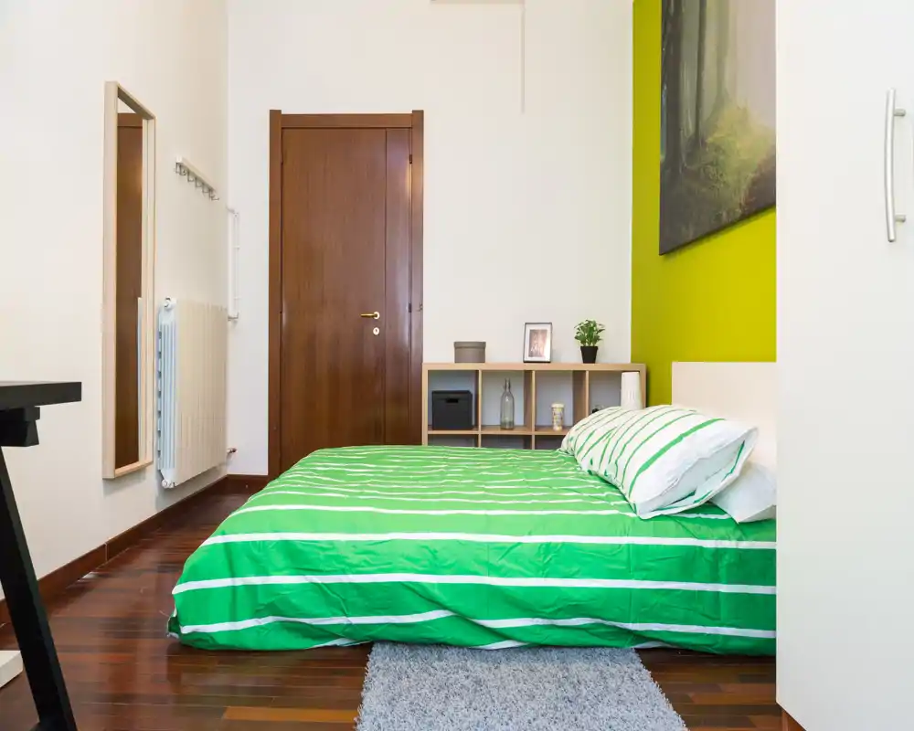 Camera da letto luminosa e curata con parete verde e biancheria a righe coordinata. Il letto è completamente visibile e la stanza presenta un piccolo scaffale, un tappeto e una decorazione essenziale.