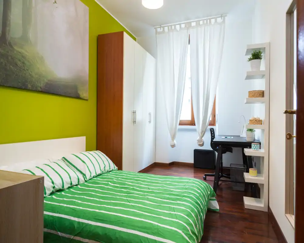 Camera da letto luminosa e moderna con parete verde, letto matrimoniale con biancheria a righe, armadio e una scrivania vicino alla finestra — ideale per soggiorni comodi o studenti.