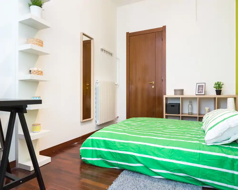 Camera da letto luminosa e ordinata con letto a righe verdi, pavimento in legno e mensole minimaliste — perfetta per annunci che mostrano uno spazio moderno e pulito.