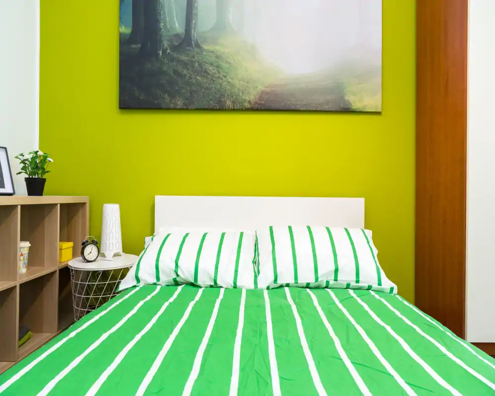 Camera da letto moderna e luminosa con copriletto a righe verdi, testiera bianca e quadro decorativo sopra. Ambiente ben illuminato e arredato con comodino, lampada e scaffale per riporre oggetti.