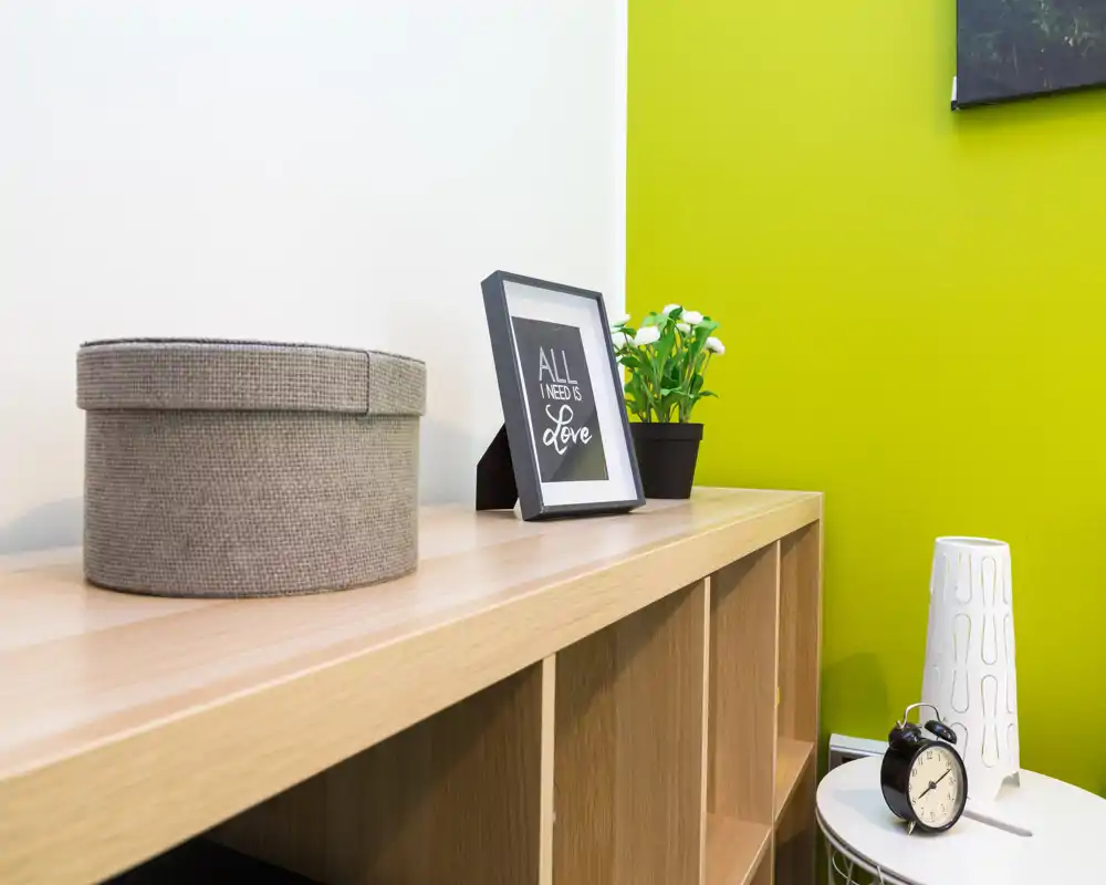 Angolo luminoso e moderno con una libreria bassa in legno, stampa decorativa incorniciata, piccolo vaso con pianta e accessori da tavolo, ideale per angoli soggiorno o lettura.