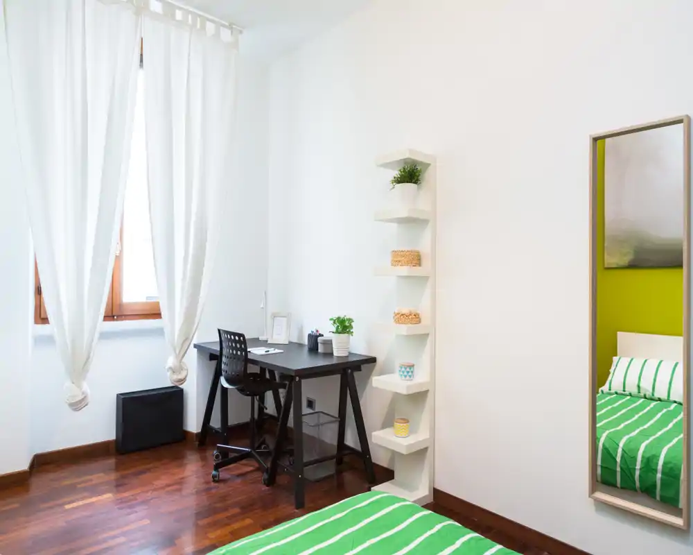 Angolo camera da letto luminoso e moderno con letto a righe verdi in primo piano, una scrivania nera vicino alla finestra con tende e scaffalatura minimale. La luce naturale e il pavimento in legno creano un ambiente fresco e accogliente, adatto a studenti o monolocali.