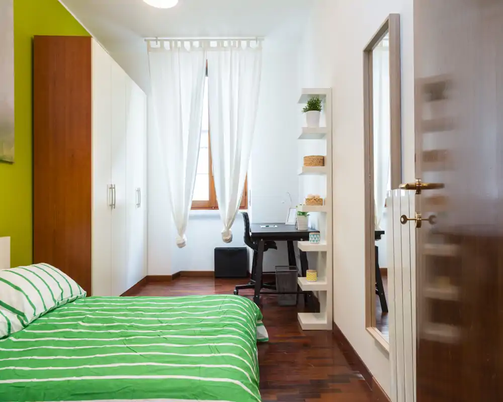 Camera da letto luminosa e curata con letto a righe verdi in primo piano, un armadio sulla parete sinistra e una scrivania vicino alla finestra — ideale per una persona singola o una stanza per gli ospiti.