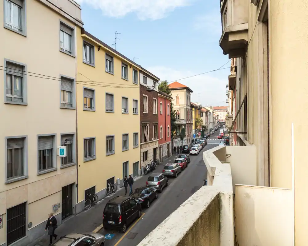 Vista sulla strada da un balcone che mostra una via residenziale con auto parcheggiate, pedoni e palazzi di medie dimensioni in pieno giorno — perfetta per mostrare il contesto del quartiere.