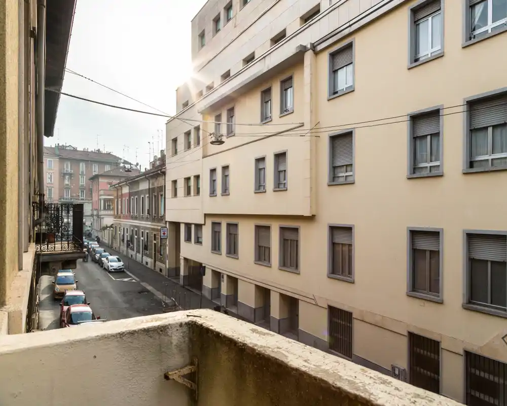 Vista dal balcone su una tranquilla strada cittadina con palazzi adiacenti e auto parcheggiate; una luce mattutina tenue illumina la scena.