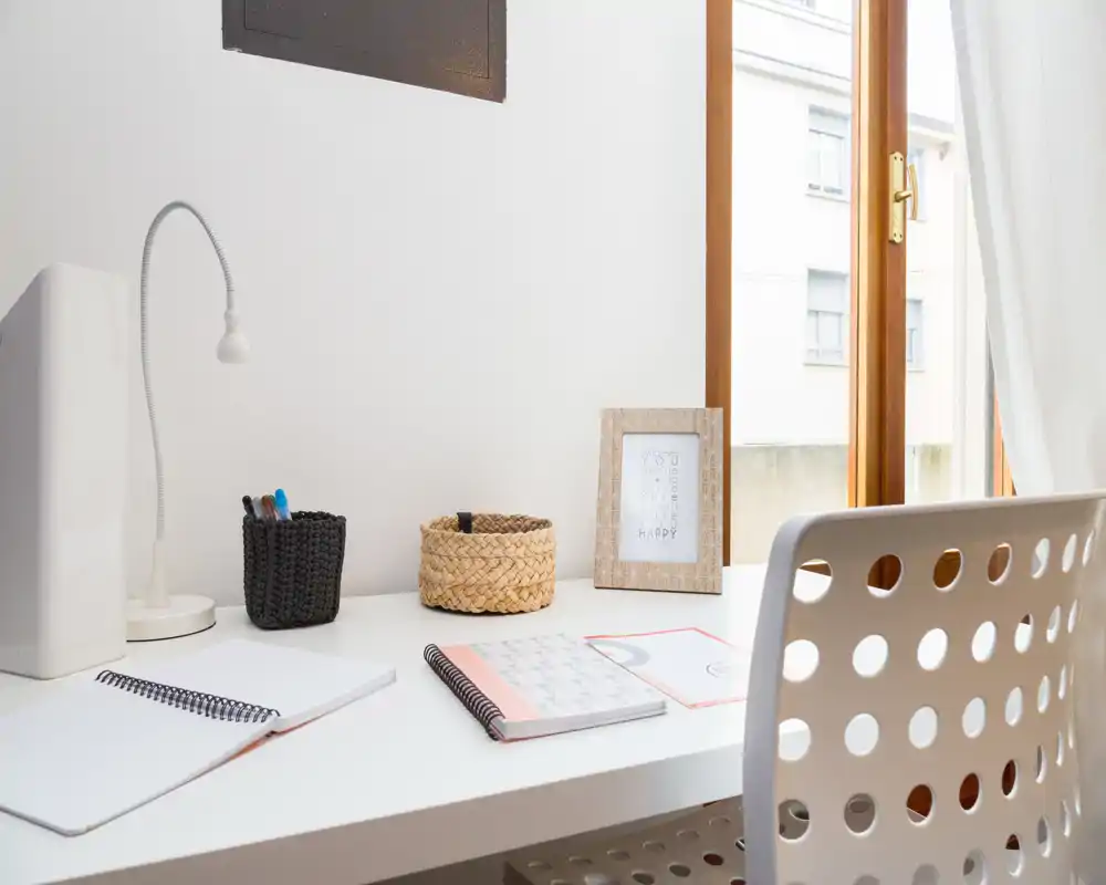 Angolo scrivania luminoso e minimal vicino a una grande finestra con sedia bianca, lampada, taccuini e cestini decorativi — un accogliente spazio per lavorare da casa.
