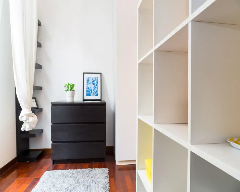 Angolo di camera minimal con comò scuro a tre cassetti, scaffalatura aperta e tappeto morbido; luce naturale intensa e dettagli semplici danno un senso moderno e pulito.