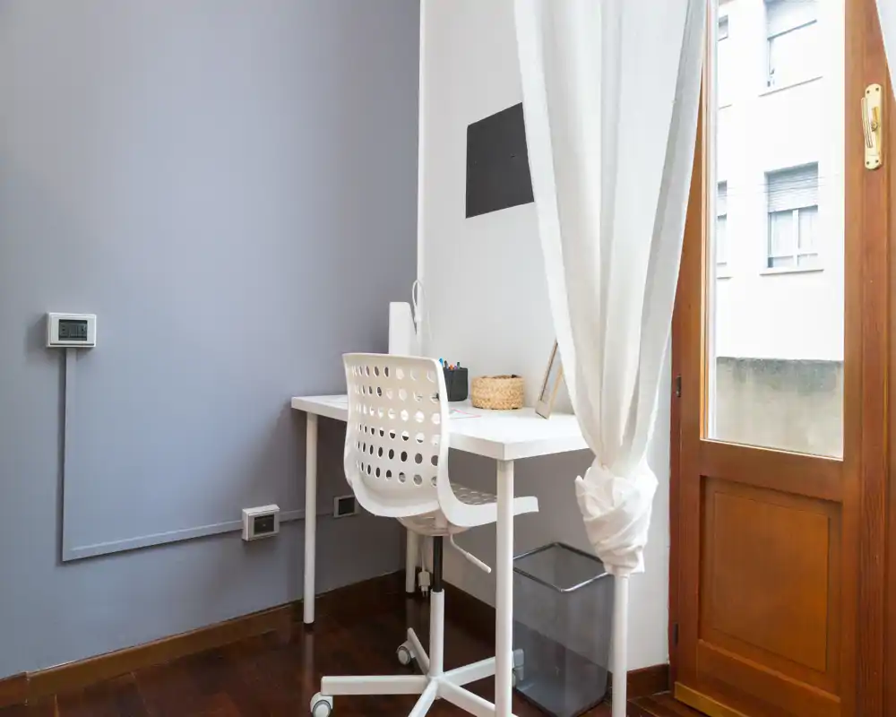 Angolo studio minimal e luminoso con scrivania bianca e sedia ergonomica accanto a una porta che lascia entrare luce naturale. Stile pulito e moderno, ideale per smart working o studio.