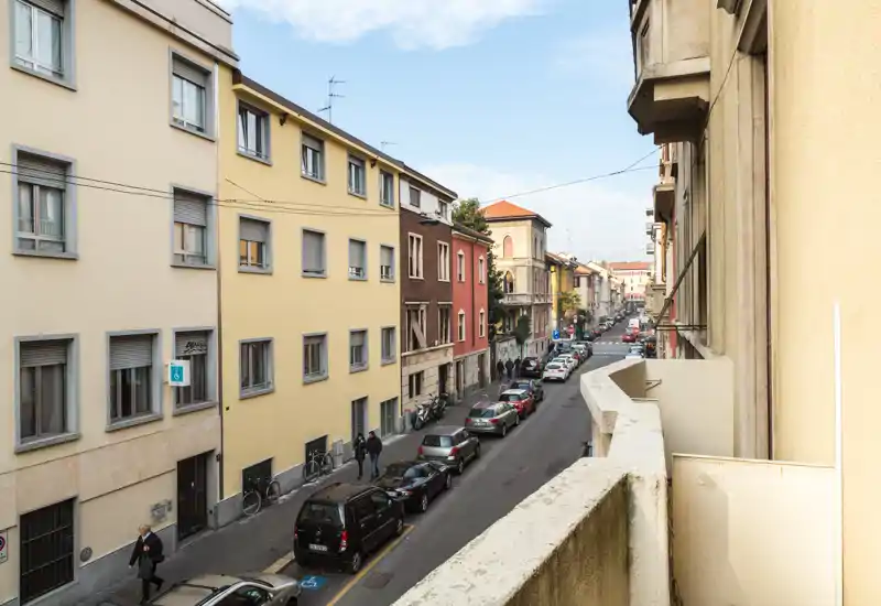 Vue de la rue depuis un balcon montrant une rue résidentielle avec des voitures garées, des piétons et des immeubles moyens en plein jour — idéale pour présenter le contexte du quartier.