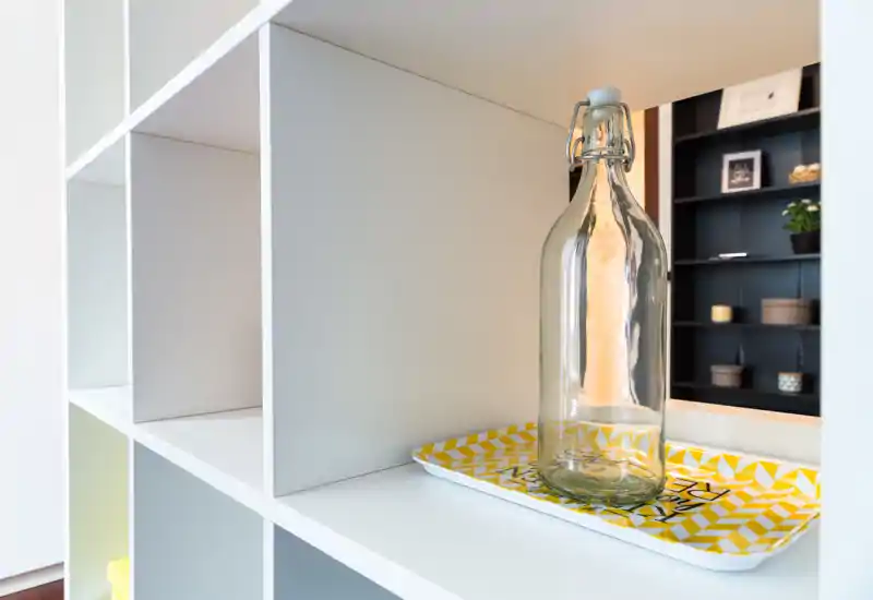 Gros plan d'une étagère modulaire blanche avec une bouteille en verre décorative sur un plateau à motifs. La composition met l'accent sur les détails de rangement et les accents décoratifs plutôt que sur une vue d'ensemble de la pièce.