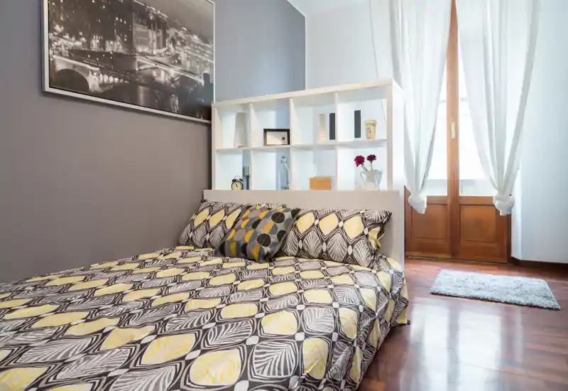Camera da letto luminosa e curata con biancheria a fantasia, cuscini decorativi e testiera a scaffale; ampia finestra con luce naturale e pavimento in legno caldo.
