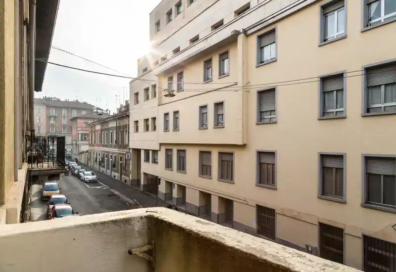 Vue du balcon sur une rue urbaine calme avec immeubles voisins et voitures garées ; une douce lumière matinale éclaire la scène.