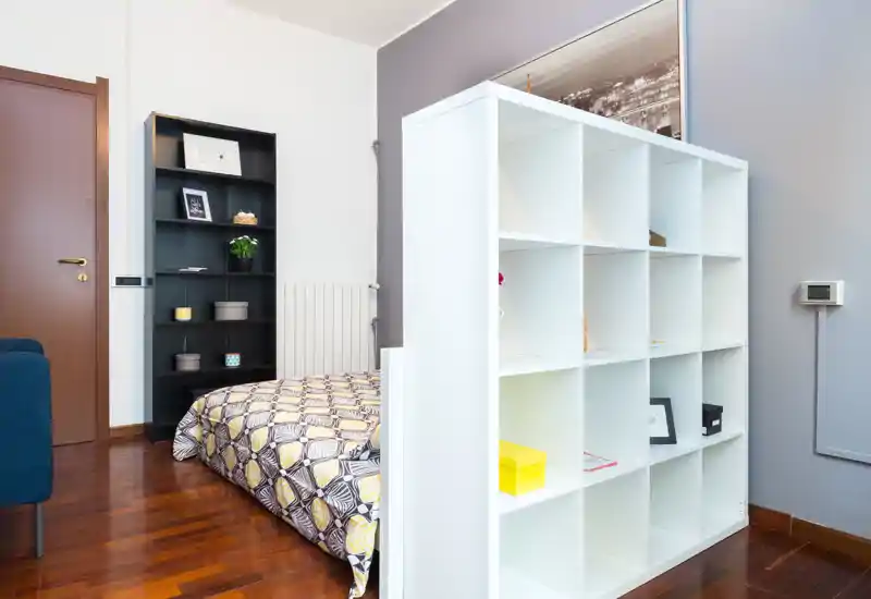 Camera da letto moderna e luminosa con un letto matrimoniale dal copriletto a fantasia parzialmente visibile dietro una libreria bianca a giorno; pavimento in legno e mensole curate creano un ambiente accogliente.
