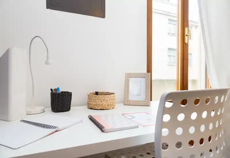 Coin bureau lumineux et minimaliste près d'une grande fenêtre avec chaise blanche, lampe, carnets et petits paniers décoratifs — un espace de télétravail confortable.