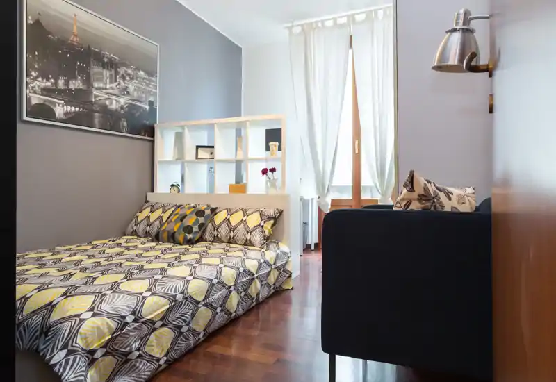 Camera da letto luminosa e moderna con copriletto decorato, letto basso e libreria a testiera; luce naturale da ampie finestre e pavimento in legno caldo creano un'atmosfera accogliente.