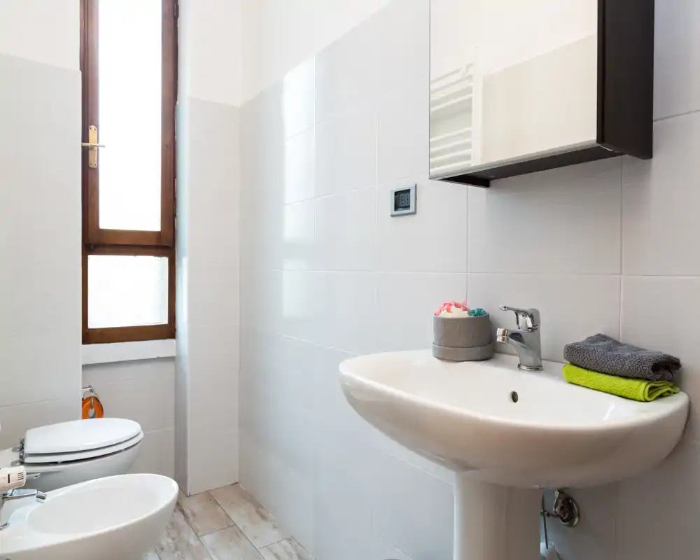 Bagno luminoso e moderno con lavabo a colonna, bidet e WC accanto a una finestra alta con vetro satinato; piastrelle chiare e pavimento effetto legno rendono l'ambiente fresco e arioso.