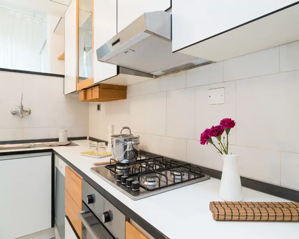 Cucina moderna e luminosa con piano cottura a gas, pentola in acciaio, lavello e piani bianchi; accenti in legno e un piccolo vaso di fiori aggiungono calore.