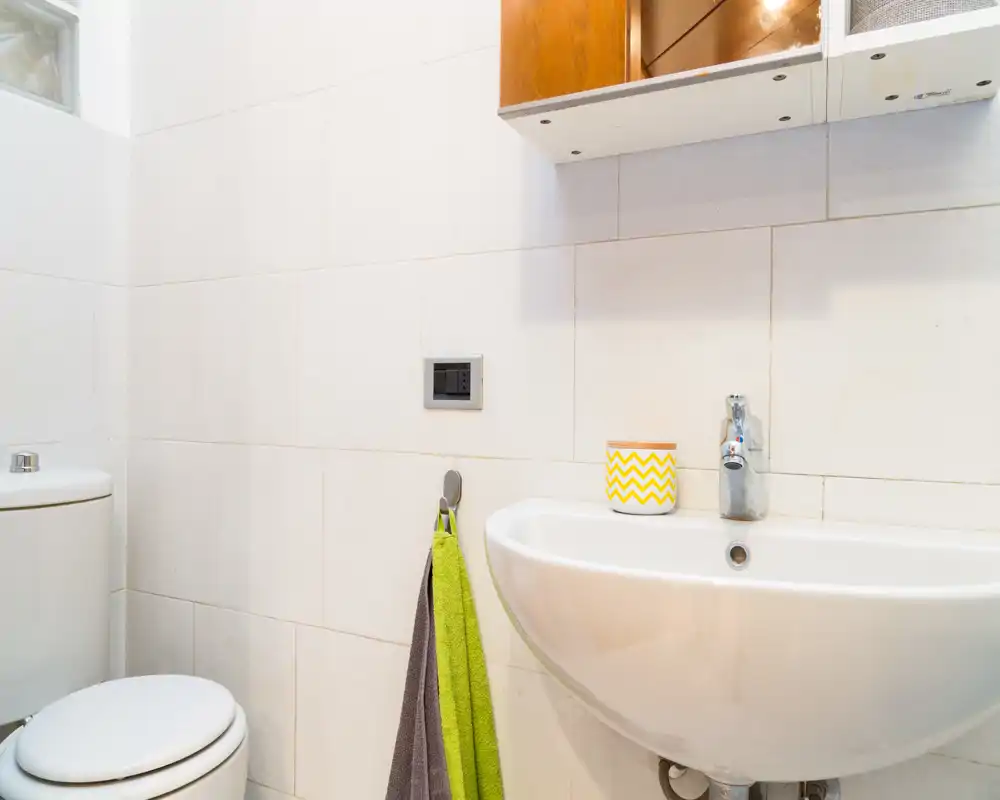Bagno compatto e pulito con lavabo bianco a parete, rubinetto cromato e WC. Piastrelle neutre e accessori semplici creano uno spazio funzionale e fresco.