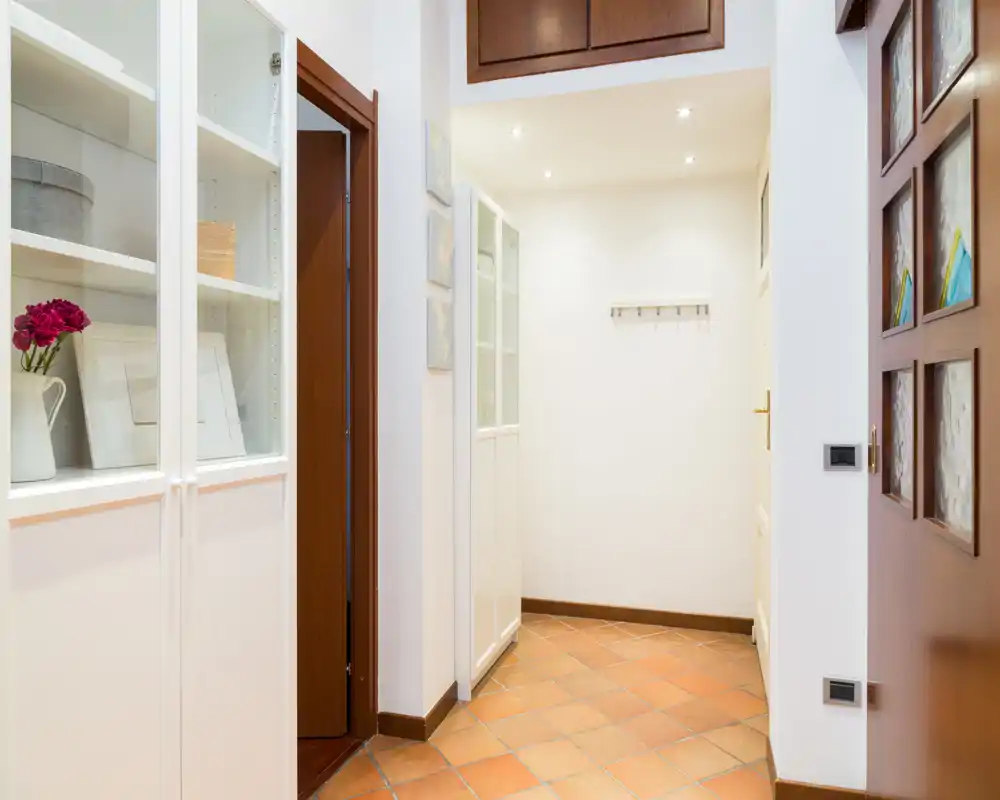 Ingresso luminoso e ordinato con pavimento in cotto, armadietti bianchi a muro e dettagli in legno caldo sulla porta — un’accogliente area d’ingresso.