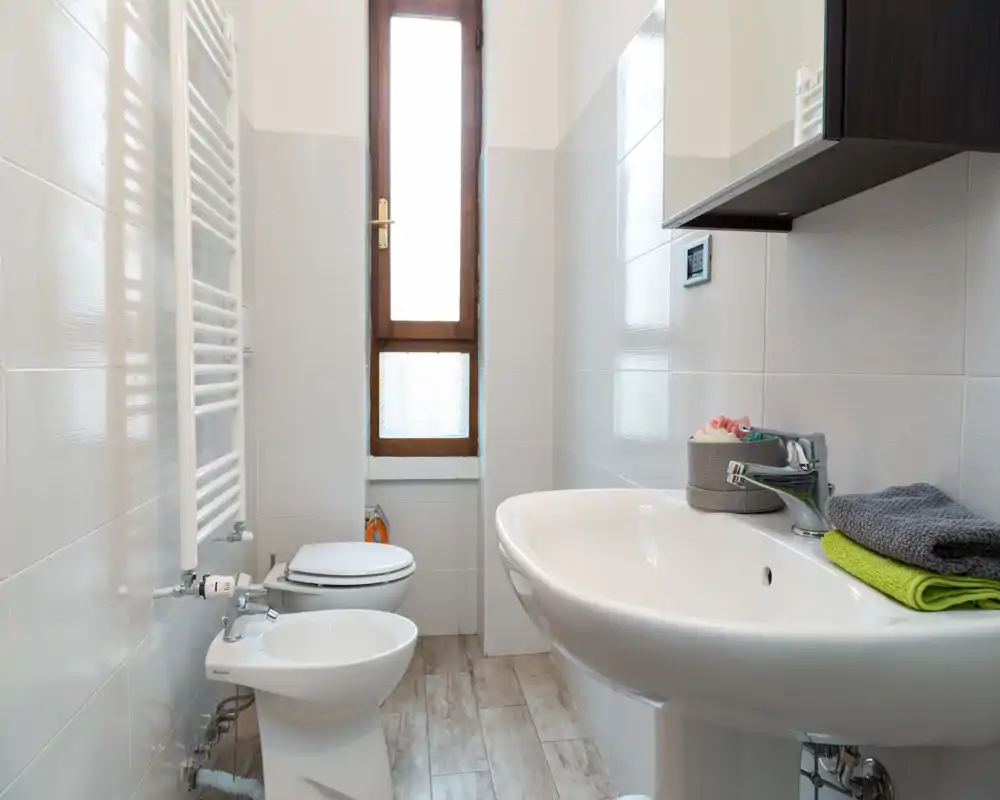 Bagno moderno e luminoso con lavabo, bidet e WC disposti in un ambiente stretto. Piastrelle bianche e una finestra alta satinata garantiscono luce naturale e un aspetto pulito.