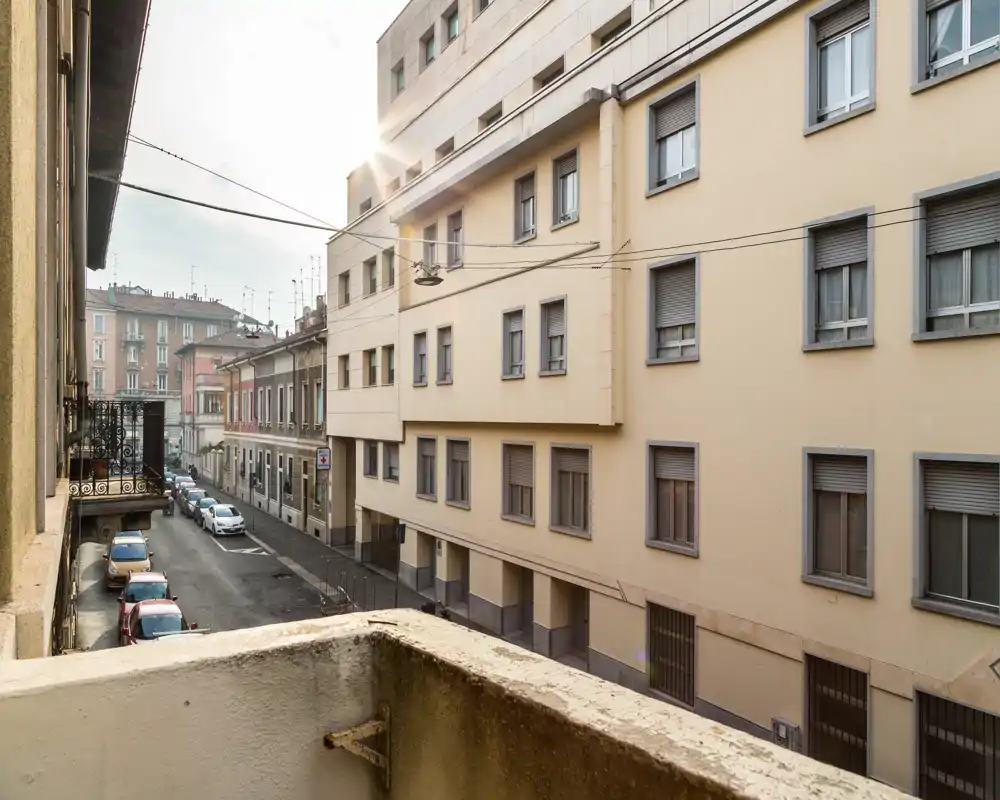 Vista soleggiata su una strada residenziale da un piccolo balcone: auto in sosta e palazzi adiacenti.