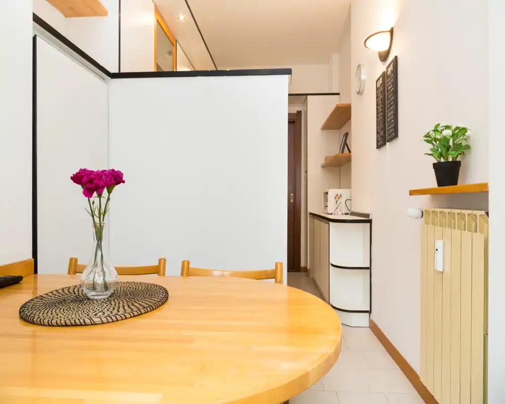 Zona pranzo luminosa e ordinata con tavolo rotondo in legno, sottopiatto decorativo e vaso di fiori; ambiente compatto che si affaccia su una piccola kitchenette.
