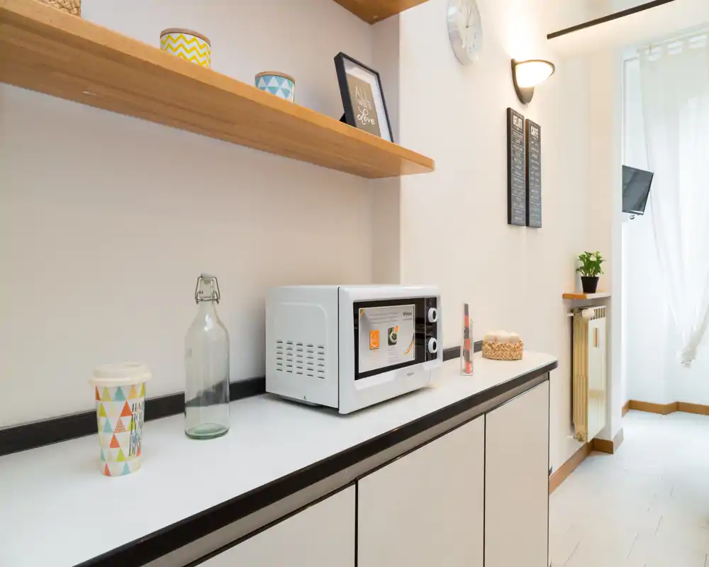 Angolo cucina compatto con microonde sul piano, tazze decorative e mensola in legno; area cucina piccola, luminosa e arredatta con gusto.