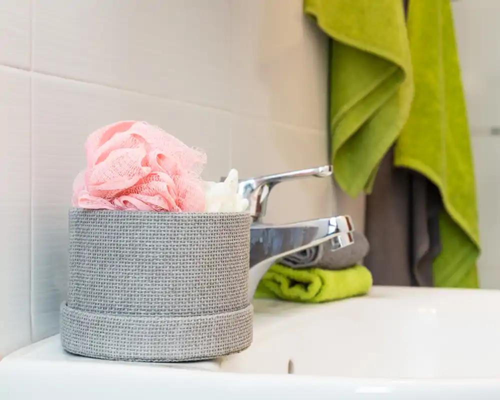 Primo piano dell'area lavabo in bagno con rubinetto cromato, contenitore testurizzato con spugne per il corpo e asciugamani piegati sul piano; immagine luminosa e curata, perfetta per annunci.