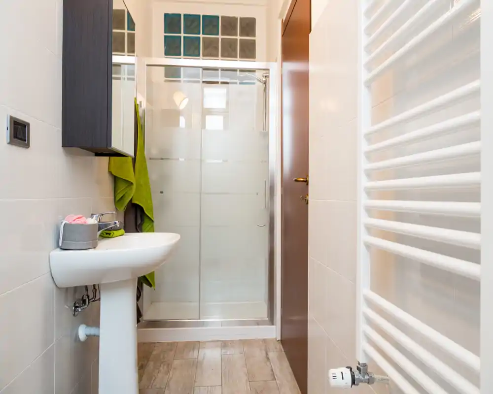 Bagno moderno e luminoso con doccia con box in vetro, lavabo a colonna e termoarredo a parete; piastrelle neutre e pavimento effetto legno per un aspetto pulito e contemporaneo.