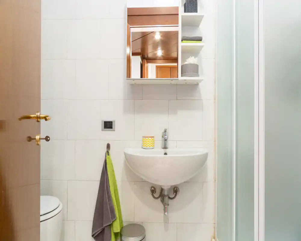 Bagno compatto e ben illuminato con lavabo a parete, pensile con specchio, box doccia con vetro satinato, appendi-asciugamani e wc parzialmente visibile. Piastrelle neutre e soluzioni contenitive semplici rendono lo spazio funzionale.