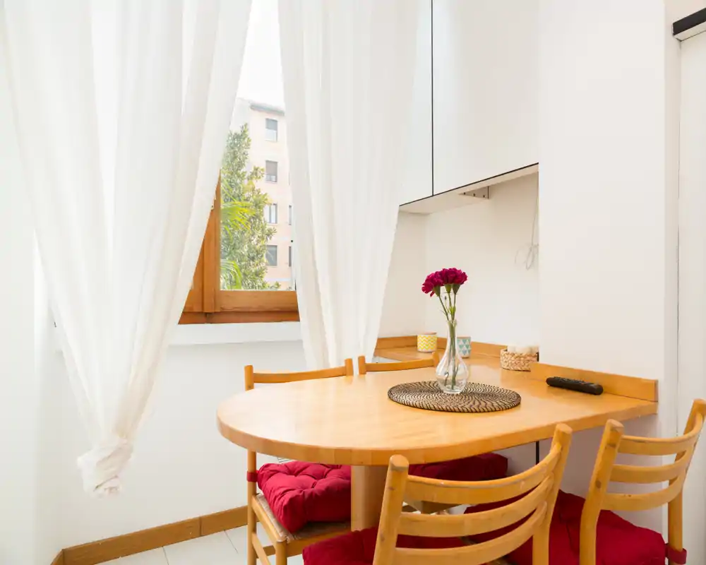 Accogliente angolo pranzo soleggiato con tavolo rotondo in legno, tre sedie imbottite e un vaso di fiori vicino a una finestra luminosa — perfetto per pasti informali.
