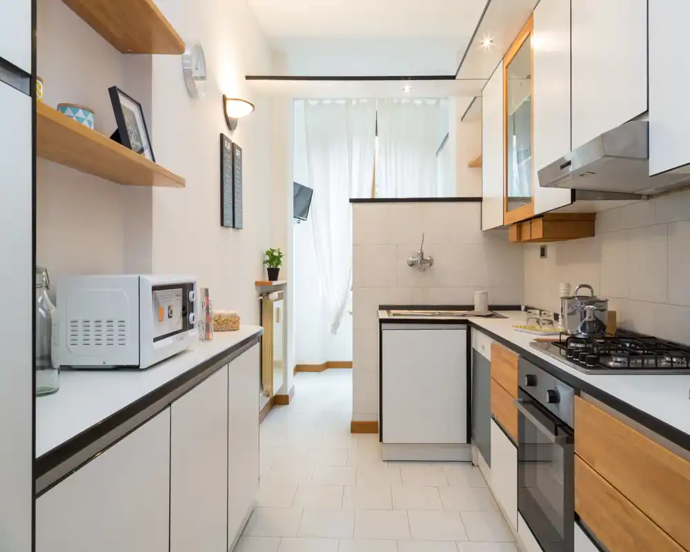 Cucina moderna a corridoio, luminosa, con ante bianche e dettagli in legno, piano cottura a gas, forno integrato e piccoli elettrodomestici ordinati.
