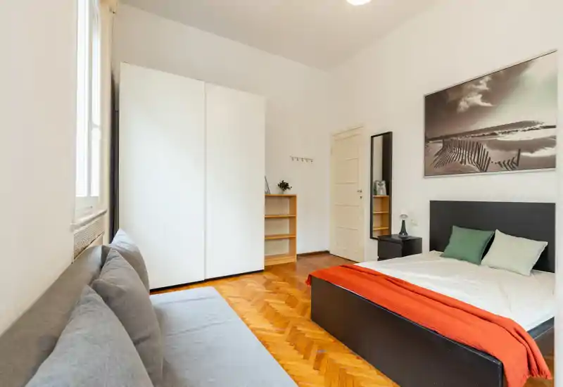 Bright, neatly staged bedroom with a double bed featuring a dark headboard, colorful cushions and an orange throw; large white wardrobe and a sofa offer extra storage and seating.